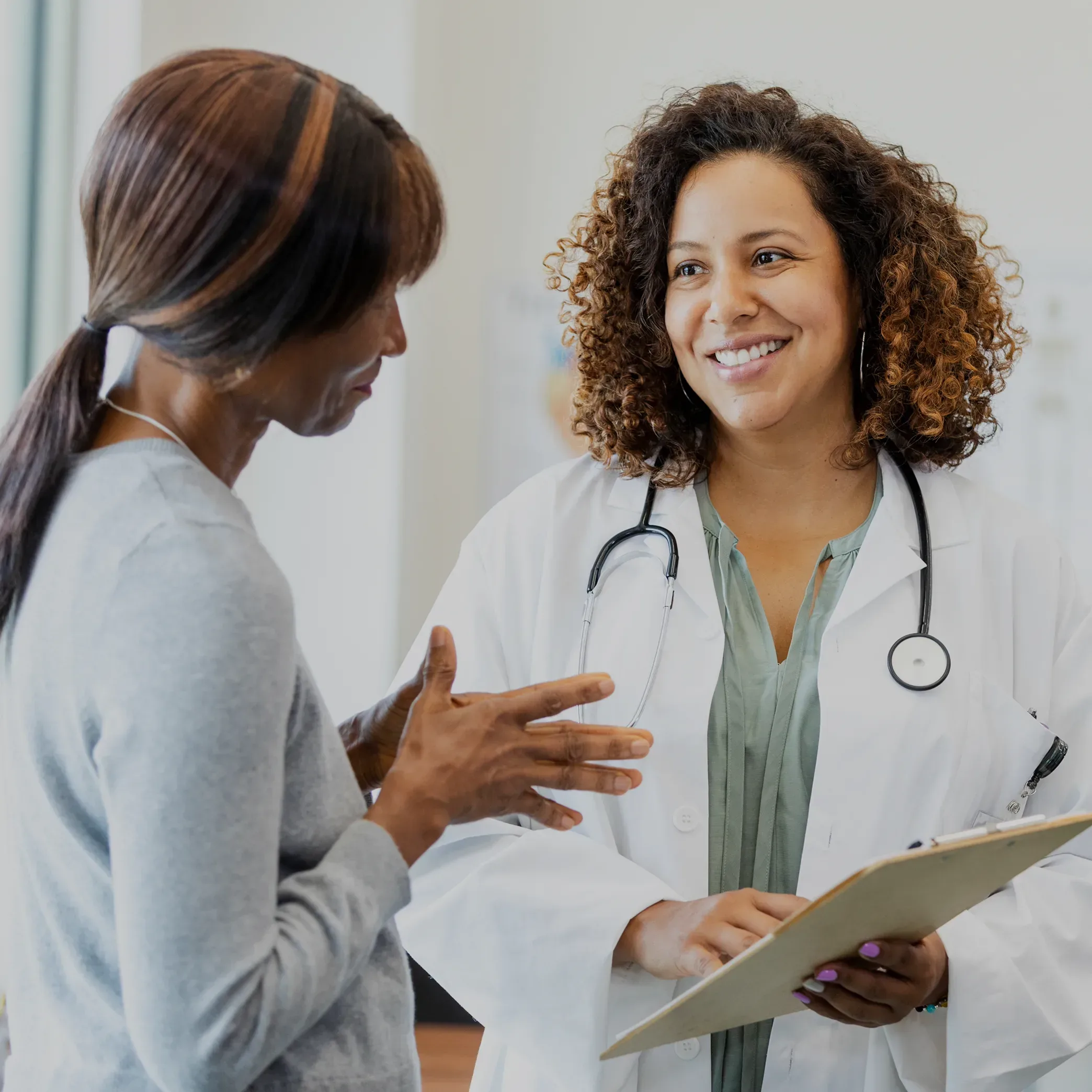 A smiling female doctor with stethoscope around her neck and clipboard in her hand consults with a patient.